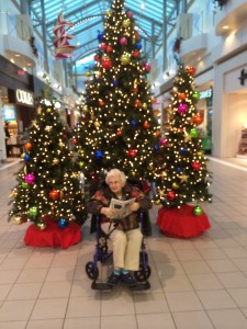 An older lady in a wheelchair sitting in front of a large christmas tree with a smaller christmas tree on each side of the large one.